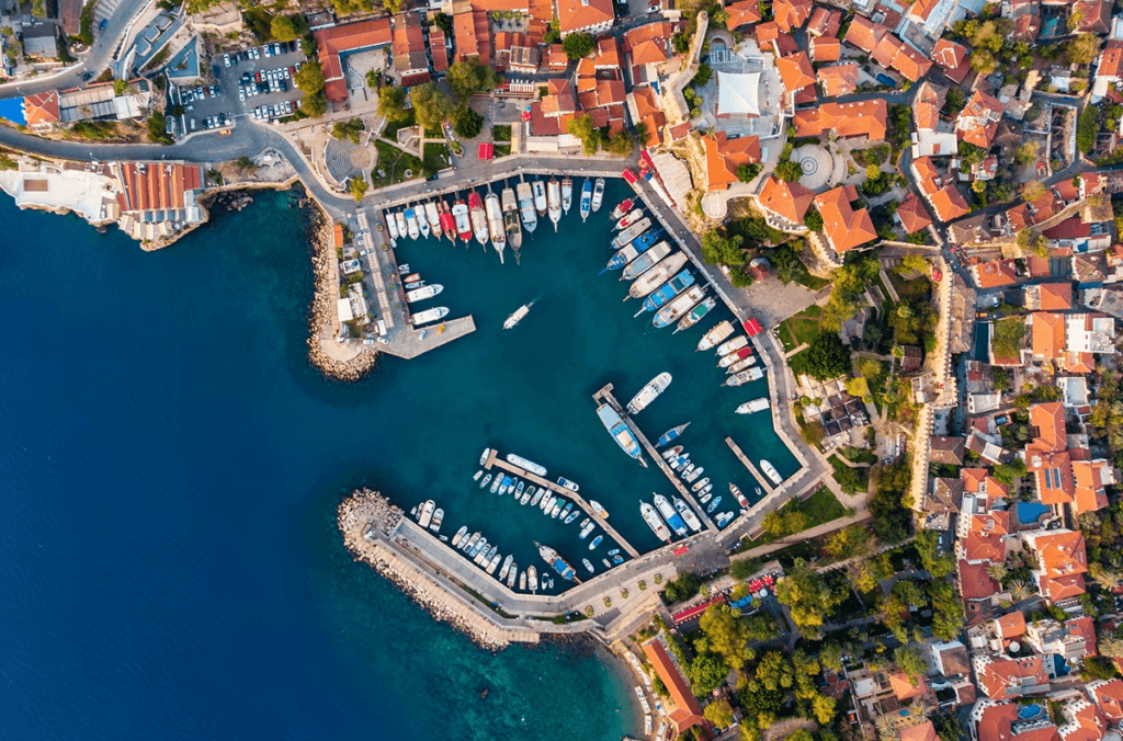 An aerial view of a coastal town with a marina filled with boats, surrounded by buildings with red-tiled roofs and lush greenery.