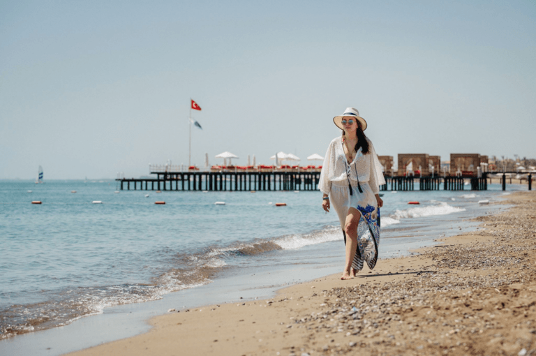 Женщина идет по пляжу с доской для серфинга под мышкой, в шляпе и солнцезащитных очках; на заднем плане — пирс и флаги.