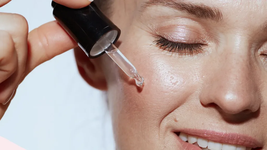 Eine Person trägt eine Flüssigkeit aus einer Pipette auf ihr Gesicht auf. Das Bild zeigt eine Nahaufnahme von Auge und Wange der Person und betont damit die Hautpflege oder Schönheitsbehandlung.