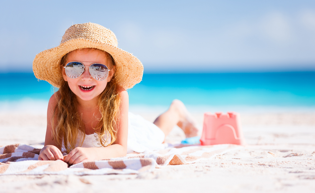 A young girl wearing a straw hat and sunglasses lies on a beach towel on the sand, smiling. There is a sandcastle and a bucket nearby, with the ocean and clear blue sky in the background.