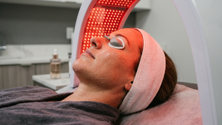 A hyper-realistic, clinical editorial portrait captured with an 85mm macro lens, showcasing a woman in profile undergoing an advanced red-light therapy treatment. The composition is intimate and focused, emphasizing the clinical serenity of a high-end med-spa. The primary light source is the intense, warm crimson glow emanating from the LED arch, which casts a dramatic, saturated flush across the subject’s face and neckline, highlighting the fine, post-treatment texture of her skin. The subject wears protective silver-toned eye goggles and a plush, soft-textured white headband that contrasts with the deep charcoal grey of her luxurious, towel-like robe. Her expression is one of tranquil, meditative stillness. The skin texture is rendered in high-definition 8k, revealing delicate, microscopic post-peel flaking and a natural, dewy luminescence reflective of the intense light. The background is softly out-of-focus, suggesting a sanitized, modern medical-aesthetic environment with clean lines and subtle product placement on a marble-top station, reinforcing a atmosphere of professional self-care and medical luxury.
